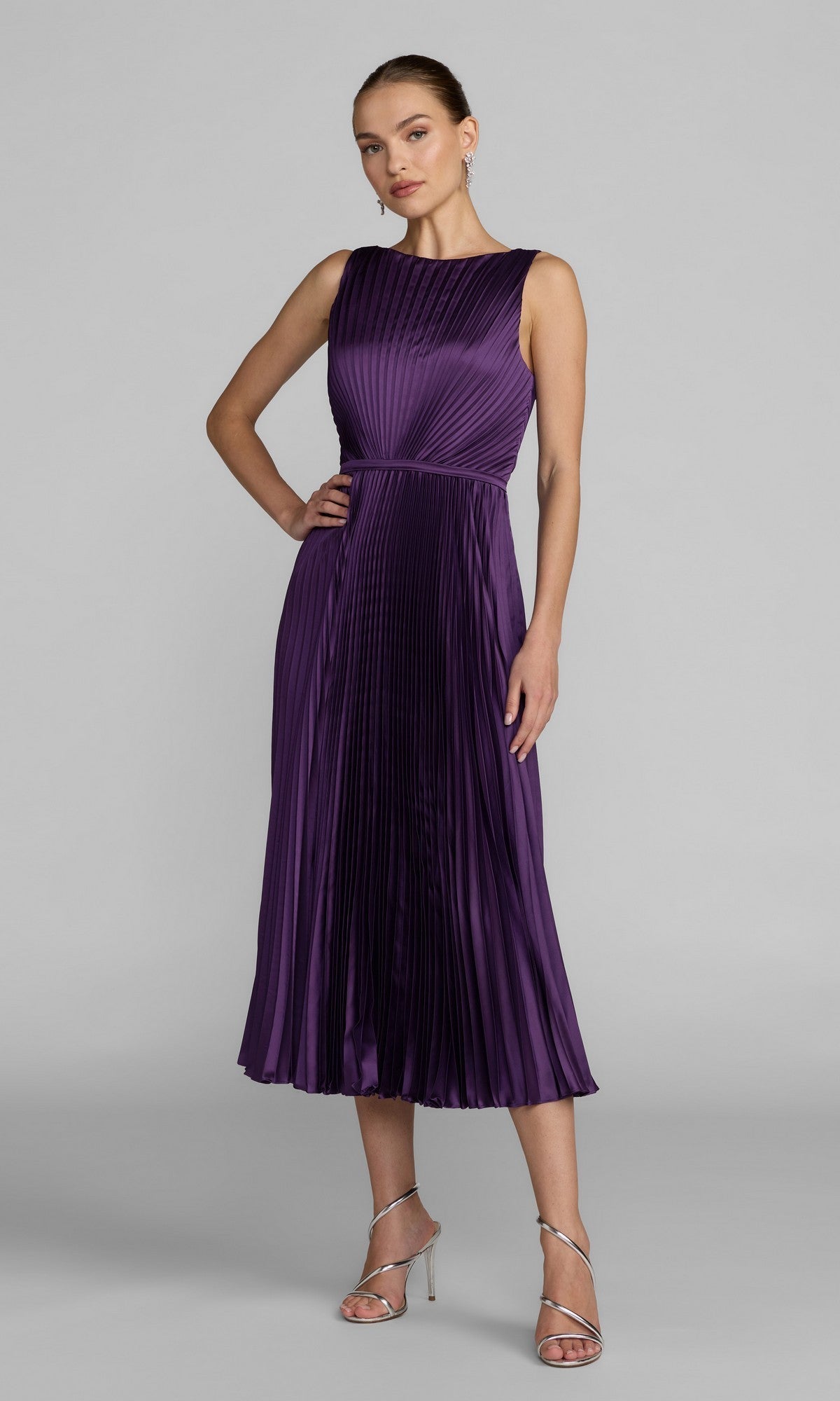A woman wearing the Mac Duggal 56326 midi party dress in purple, featuring a sleeveless, pleated charmeuse bodice and an elegant A-line skirt, paired with silver strappy heels and standing against a plain light gray background.